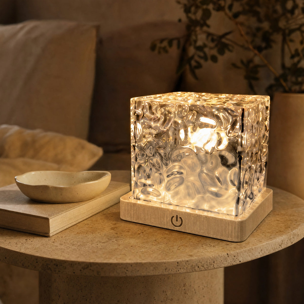 Cube lamp glowing warm amber on a travertine side table beside a linen book and ceramic dish