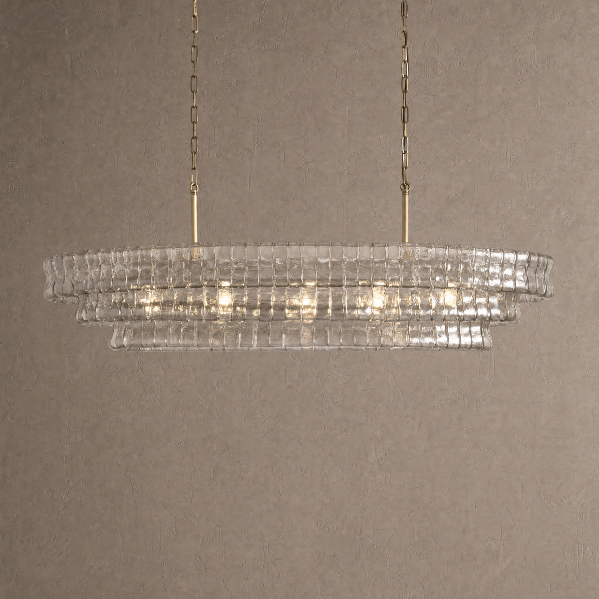 Ghiaccio oval chandelier in clear ribbed hand-blown glass with twin antique brass chains against a warm neutral wall
